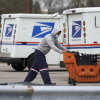 A U.S. Postal Service employee wearing dark blue pants, a gray hoodie and a light blue medical mask pushes a large orange plastic cart in a parking area outside a post office in Wheeling, Illinois, in 2021. Two Postal Service delivery trucks are in the background.