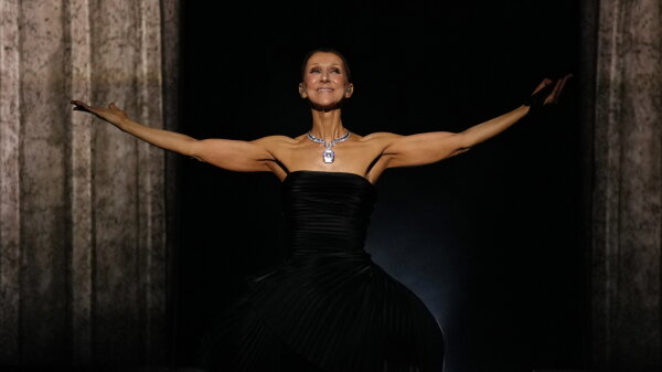 Singer Celine Dion on stage with her arms outstretched. She is wearing a black dress. 