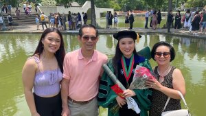 NhuNgoc Pham is shown in a cap and gown, signifying her doctorate. She holds flowers and is standing in front of a fountain with her family. 