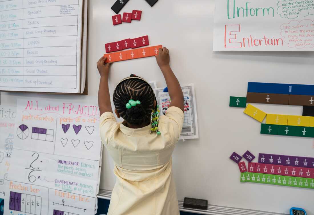 Photographed with her back to the camera, Aura places fraction blocks on the whiteboard in her third-grade class.