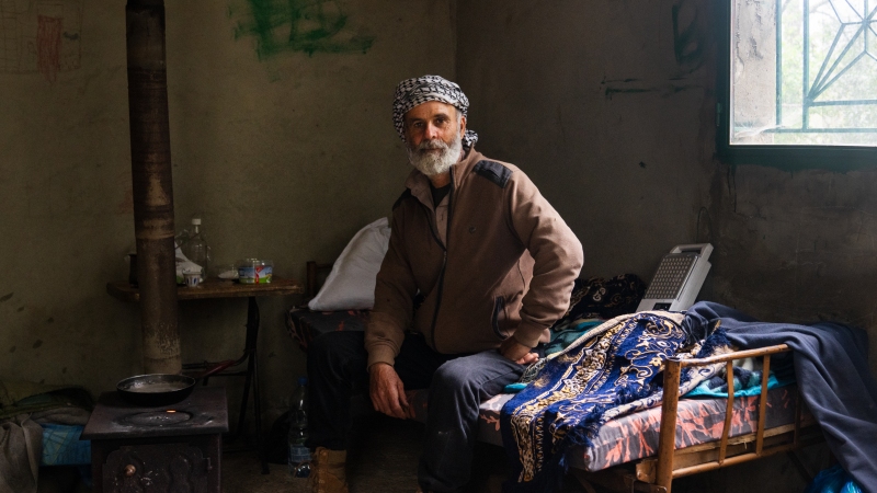 Joseph Elias Issa, 56, sits on a day bed next to a wood burning stove in a small cement building. He has a white beard and a scarf wrapped on his head.