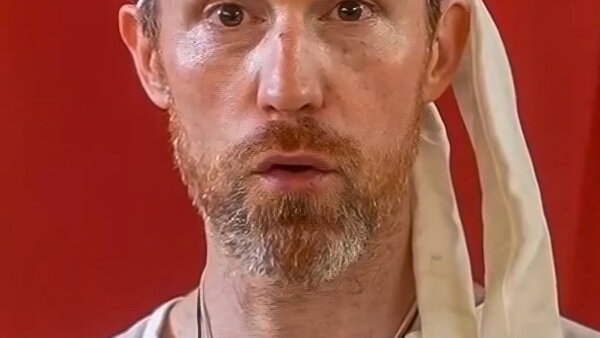 A close-up of a man's face with a red and silver-haired close-cut beard. There is a bright red background. 