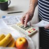For square image: A white counter with a plastic bag containing an apple and a bunch of bananas, there is an elderly light skinned woman holding a receipt in her left hand and typing numbers into an old calculator with her right hand. She is wearing a black and white striped shirt and there is a potted plant and a coffee mug in the background.