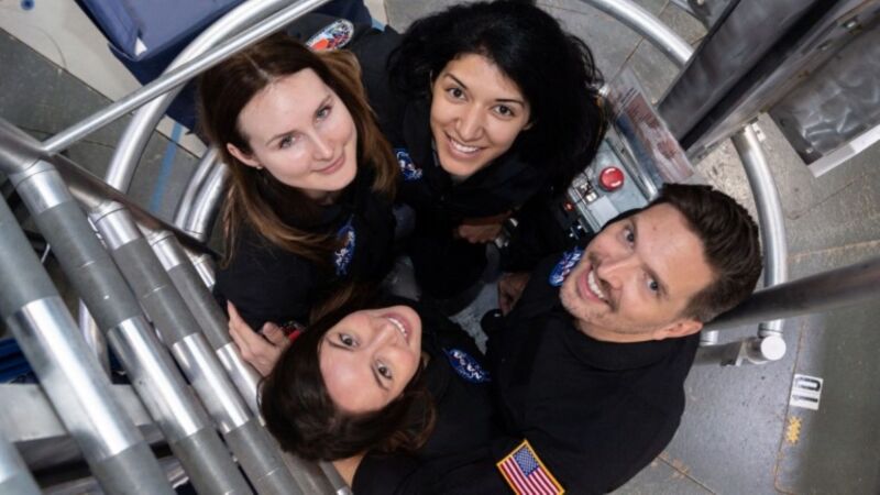 Four people smile up at the camera. They are wearing dark uniforms and one person has a U.S. flag patched onto the sleeve. They are close together, in a small, metallic, well-lit ladder well.