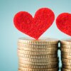 Two red felt hearts balance atop two separate stacks of gold coins against a light blue backdrop, representing people in a romantic relationship merging finances.