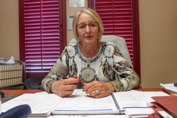 Debbie Cox, 65, has blond hair and wears glasses. She sits behind a desk and has a concerned expression on her face. 