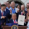 President Donald Trump is shown proudly holding up a piece of paper that shows his distinctive large, Sharpie-penned signature. He is surrounded by many other men and one woman. 