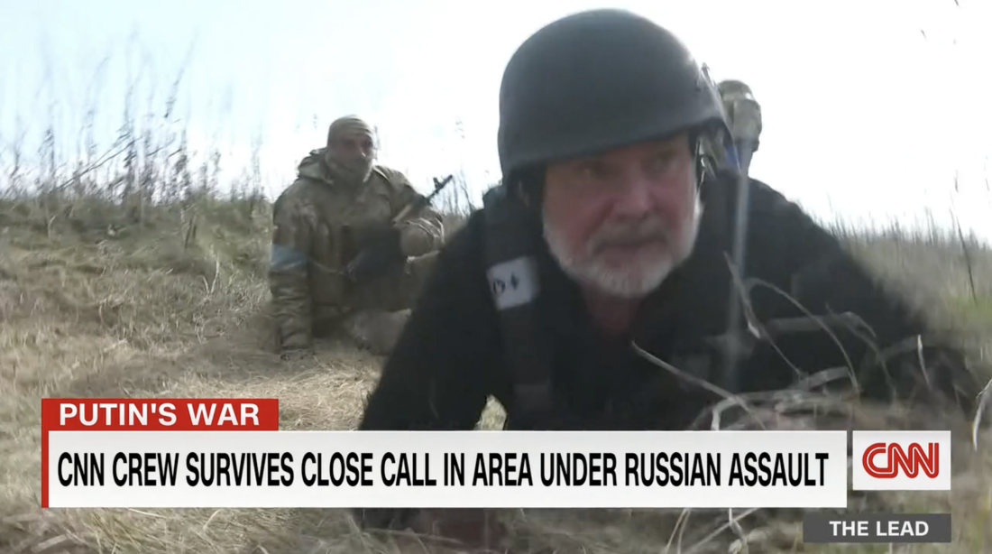 CNN's Ben Wedeman, who's wearing a helmet, ducks to the ground for cover while reporting in Ukraine in 2022. A soldier with a firearm sits on the ground behind him.
