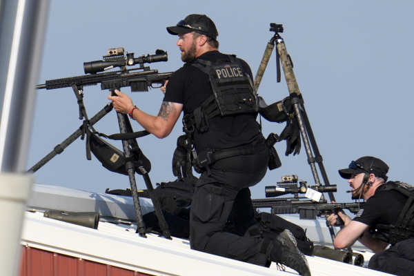 Two male police snipers positioned on a roof return fire after shots were fired while former President Donald Trump was speaking at the campaign rally on July 13. The snipers are wearing all-black uniforms and are aiming their weapons.