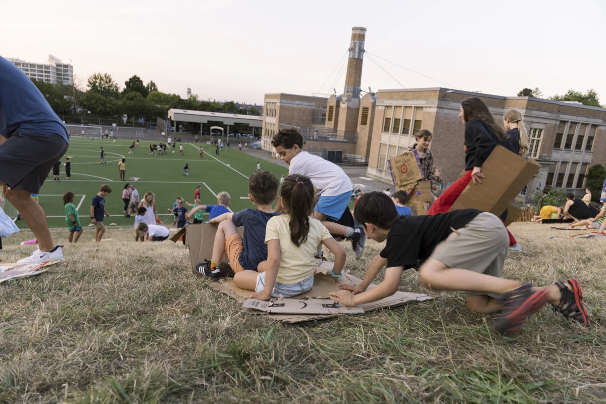 Kids slide down a grassy hill on scraps of cardboard while waiting for the Vaux’s Swifts to gather at sunset in Portland, Oregon.