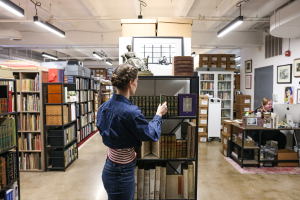 Rare book collector Rebecca Romney arranges novels at Type Punch Matrix. Rows of bookshelves are seen behind her and a person works at a computer, surrounded by books and cardboard boxes.