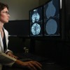 Dr Olena Weaver, wearing a white layer and glasses, looks at two mammography images on a computer screen. The one on the left has many silver structures; The other is mainly dark gray with only a few light points.