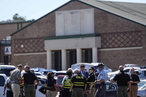 This photo shows law enforcement officers and first responders responding to Apalachee High School in Winder, Ga., on Wednesday, after a shooting was reported. Multiple adults, some wearing vests saying "GBI" and "SHERIFF" on the back, are standing outside the school. Parked cars are in the foreground.