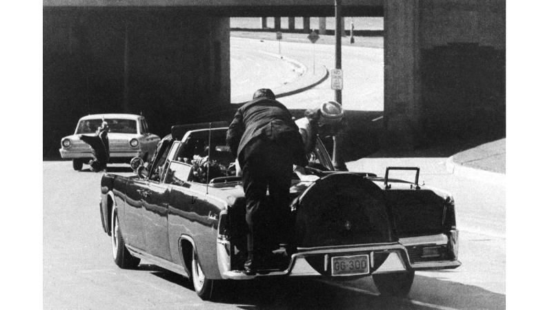 President John F. Kennedy slumps down in the back seat of the presidential limousine as it speeds along Elm Street toward the Stemmons Freeway overpass in Dallas, Texas, after being fatally shot, Nov. 22, 1963. First lady Jacqueline Kennedy leans over the president as Secret Service Agent Clint Hill pushes her back to her seat.