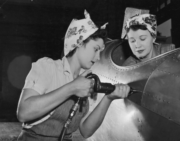 circa 1943: One American female worker drives rivets into an aircraft while another sits in the cockpit on the US home front during World War II. They wear aprons and their hair tucked into scarves. Women who went to work in industries to aid the war effort became known under the moniker 'Rosie the Riveter'.
