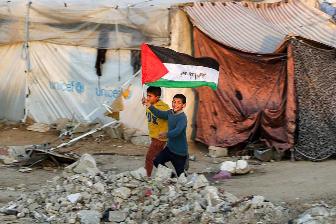 In this photo, two boys, one holding a Palestinian flag, run past tents at a camp for people displaced by conflict in Gaza. One tent is made from a white tarp displaying UNICEF's light blue logo. Concrete-looking rubble is piled on the ground.