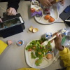 Photographed from above, seventh-graders sit together at a cafeteria table during lunch at a school in Brooklyn, New York, on February 10, 2023. One student is eating a salad, and another has a plate with tortilla chips and fruit on it. Two tablet devices and multiple cartons of milk are also on the table.