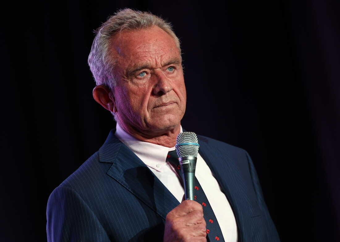 RFK Jr., wearing a suit and holding a microphone, stands against a black backdrop.