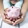 This photo shows a child's outstretched hands holding a pink piggy bank. The outstretched hands of an adult who's behind the child are cupping the outside of the child's hands.