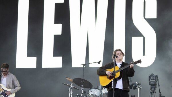  Lewis Capaldi performs at the Glastonbury Festival at Worthy Farm in Somerset, England, on June 27, 2025. He holds an acoustic guitar as he stands at a mic. The name ‘Lewis’ is seen behind him in large capital letters.