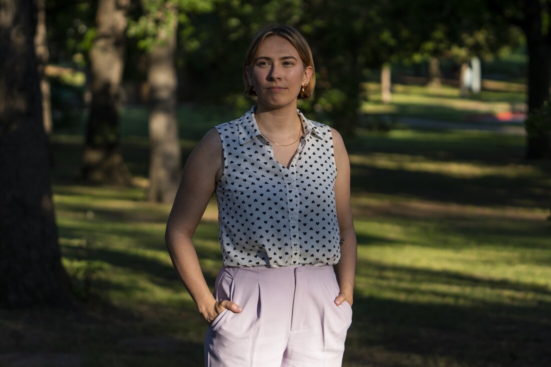 Caitlyn Mai poses for a portrait in a park near her home in Bethany, Okla., on May 18, 2024. She's wearing light-colored pants and a sleeveless white blouse with small dark hearts on it.