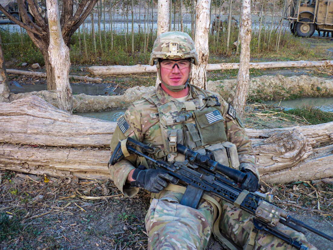 Dustin Ingram is photographed in uniform holding a large rifle in while sitting on the ground in eastern Afghanistan.