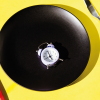 Studio photograph shows a large, black, ceramic dinner plate on a yellow background. A metal fork and knife with red plastic handles are on the right and left of the plate and a a small, white alarm clock rests on the black plate.
