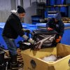 Two men at the Portland Fish Exchange in Portland, Maine, load large silver fish from a black plastic crate into a large yellow plastic tote box.