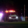 Neon lights on emergency vehicles flash in the dark. On the right of the photo, a security officer raises the boom barrier.
