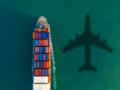 This aerial photo shows a cargo ship loaded with colorful shipping containers and floating on greenish-blue water. A shadow of an airplane is on the surface of the water, to the right of the ship.