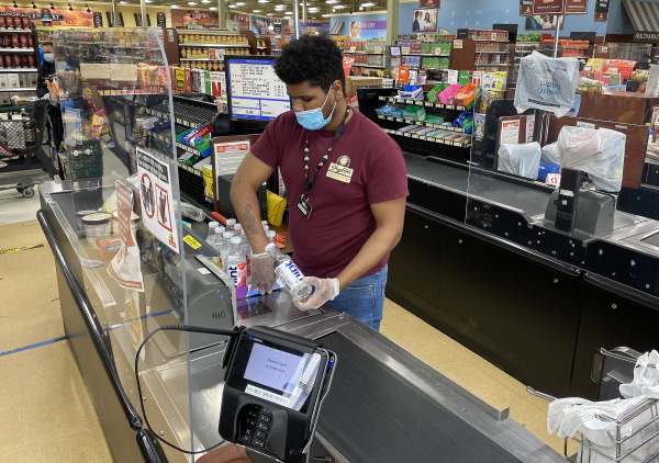 This photo shows a grocery store checkout clerk working behind a plexiglass divider at the ShopRite supermarket in Uniondale, N.Y., at the start of the coronavirus pandemic. He's wearing a light blue medical-style mask over his mouth and is scanning bottled beverages that are on a black conveyor belt in the checkout lane. Aisles of food stand behind him in the background.