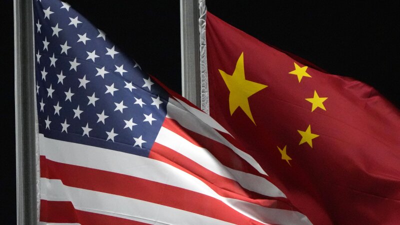 In this photo, a U.S. flag is on the left, and a Chinese flag is on the right. Both are attached to flagpoles, and they wave against a black night sky.