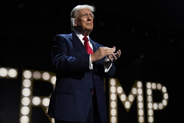Republican presidential candidate Donald Trump is introduced during the final night of the Republican National Convention. He's standing on a stage while wearing a dark suit and red tie. A bandage is on his right ear, and he's clapping his hands. Behind him, lights spell out his last name.