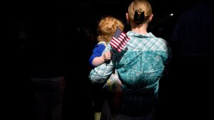 A young girl holding an American flag is held by her mother. Both face away from the camera.