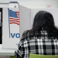 A person with their back to the camera marks their ballot in a voting booth bearing a U.S. flag and the word "Vote."