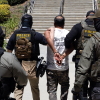 Four Immigration and Customs Enforcement agents walk a detainee out of the Ventura County Government Center in Ventura, California, on July 27. The individuals are photographed with their backs to the camera, and the detainee is handcuffed behind his back. The agents are wearing bulletproof vests that say "POLICE ICE" in yellow capital letters.