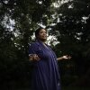 Wearing a dark blue dress, Farah Larrieux stands in her backyard in South Florida. Multiple trees stand in the background.