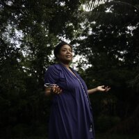 Wearing a dark blue dress, Farah Larrieux stands in her backyard in South Florida. Multiple trees stand in the background.