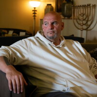 Senator John Fetterman sits in a room in Jerusalem with a menorah in the background. Fetterman is wearing a white hooded sweatshirt and looking off camera. 