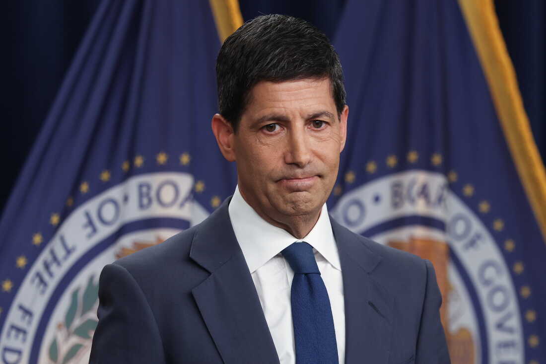 This photo shows Federal Reserve Chairman Kevin Warsh. He's wearing a blue suit, white shirt and blue tie. Behind him are two flags with the seal of the Federal Reserve on them.
