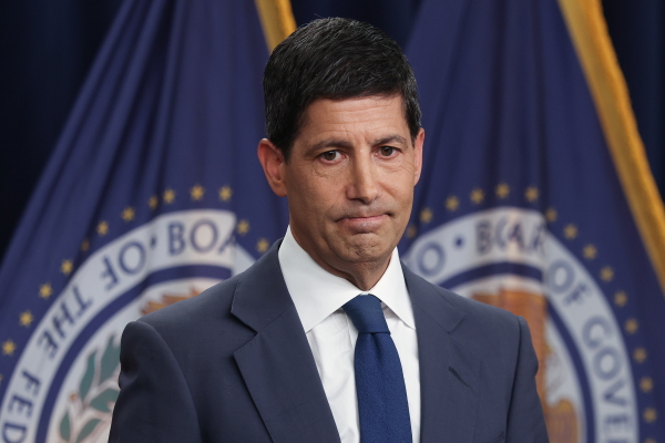 This photo shows Federal Reserve Chairman Kevin Warsh. He's wearing a blue suit, white shirt and blue tie. Behind him are two flags with the seal of the Federal Reserve on them.