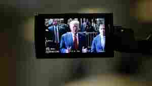 This photo shows former President Donald Trump on a camera monitor as he speaks to the press in February outside Manhattan Criminal Court. He's wearing a blue suit jacket, a red tie and a white shirt. Many people in suits and law enforcement uniforms stand behind him. 