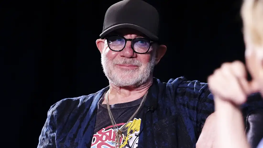 Bernie Taupin smiles. He wears a dark blue lightweight jacket over a graphic tee and a black cap. He has a gray close-cut beard. He is seen from the chest up.