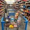 This photo shows a shopping cart in a store aisle filled with groceries. The cart is filled with items including bagels, grapes, salmon and juice. The shelves lining the aisle are filled with bags of potato chips, candy and other packaged nonperishable foods.
