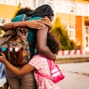 Photograph of a mother embracing her two daughters at school drop off. The school-aged children wear backpacks and are seen in front of their school building. The family is pictured from behind. Talking through what to expect at school before a new year begins and adopting a goodbye ritual are two tips from experts on helping to prepare your child for the changes as they begin a new year.