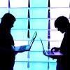 Deux hommes se détachent sur un fond bleu et violet illuminé. Ils se font face, mais regardent les ordinateurs portables qu’ils tiennent dans leurs mains. 