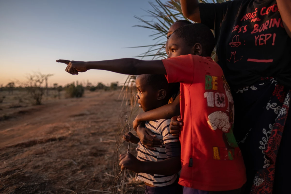 Children stand at the broken fenceline of Mosi-oa-Tunya National Park at dusk and watch as animals begin to cross into the outskirts of Livingstone, Zambia.