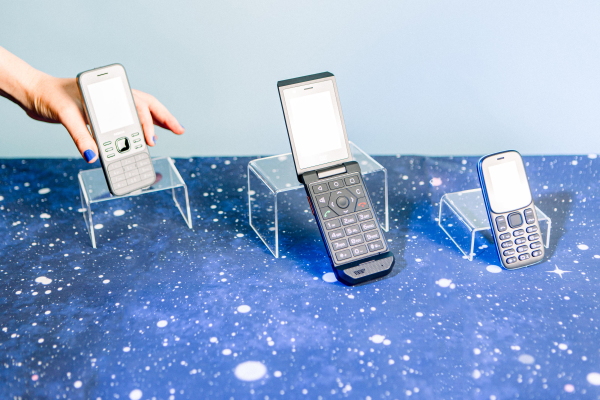 A studio photograph of three basic phones, the Nokia 6300 4G, the Cat S22 Flip and the BLU Z5 -GSM, propped up on clear display shelves. They sit on a table covered with celestial-patterned cloth and behind them is a light blue backdrop. A white hand with blue finger nails reaches in from the top left corner to take the Nokia 6300 4G phone.