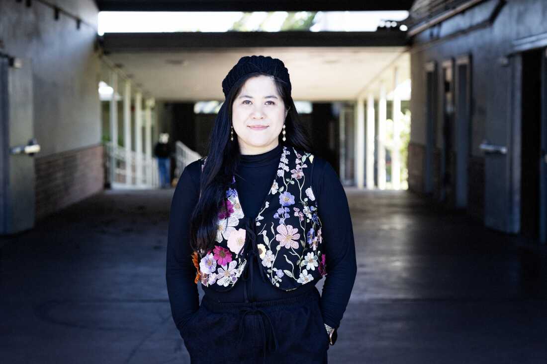 Tamamen siyah, çiçekli yelek ve örgü bere giyen genç bir Filipinli kadın olan Mary Acebu, Riverview Ortaokulu'nun açık hava girişinin ortasında duruyor.