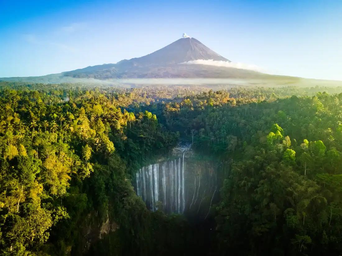The stunning Tumpak Sewu cascades like liquid silver from the cliffs, with the powerful Senaru Volcano rising in the distance, sending smoke into the sky as the landscape unfolds in all its natural glory.